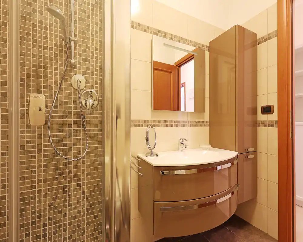 Baño moderno y luminoso con mampara de ducha de vidrio, pared de mosaico y mueble lavabo curvo con espejo.