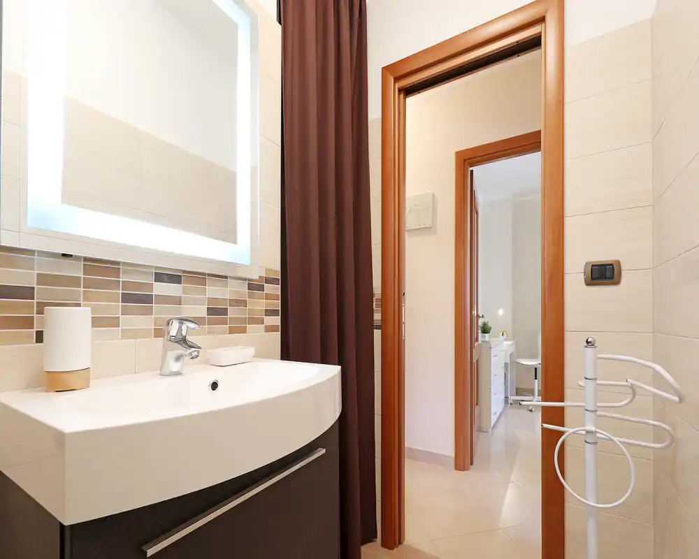 Baño moderno y luminoso con lavabo mural, espejo iluminado y alicatado decorativo. El espacio está limpio y bien iluminado, mostrando el área del lavabo y la puerta hacia la habitación contigua.