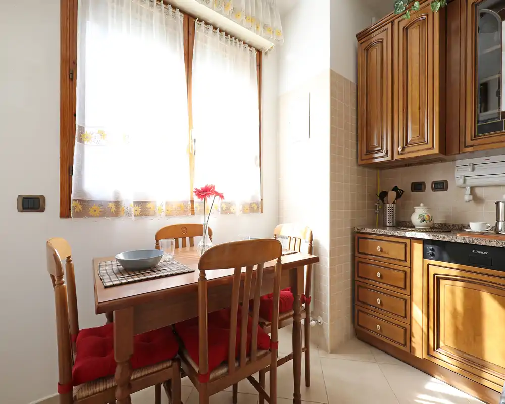 Cocina luminosa con muebles de madera y una pequeña mesa para cuatro; tonos cálidos, luz natural y cojines rojos que crean un rincón desayuno acogedor.