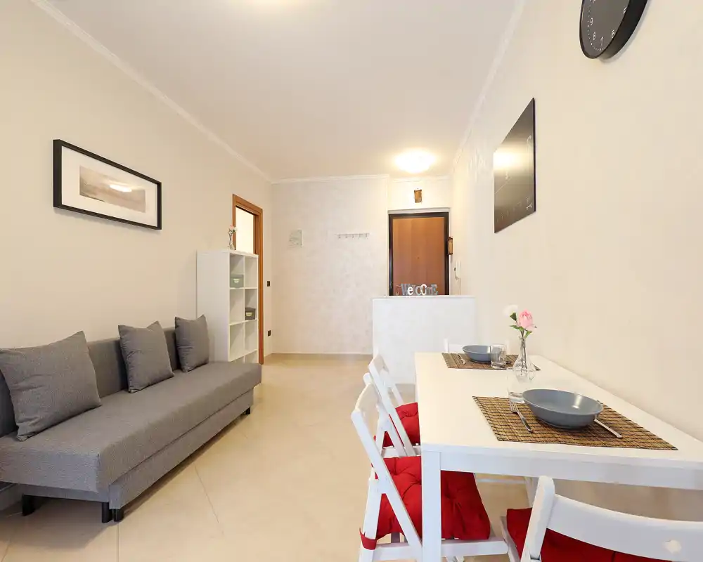 Zona de estar moderna y luminosa con sofá gris, cojines decorativos y mesa de comedor para dos. Tonos neutros y distribución ordenada crean un ambiente cálido y acogedor, ideal para parejas o grupos pequeños.