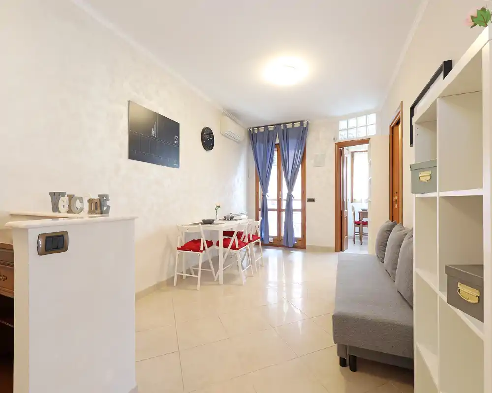 Sala luminosa y bien decorada con mesa de comedor compacta, sofá gris y estanterías — ideal para un piso urbano. La luz natural por la puerta del balcón aporta sensación de amplitud y calidez.