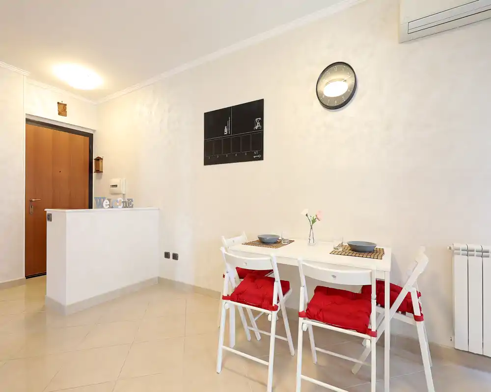 Pequeña zona de comedor con mesa blanca y cuatro sillas plegables con cojines rojos, servicio sencillo y un pequeño jarrón: luminosa, ordenada y apta para un apartamento pequeño.
