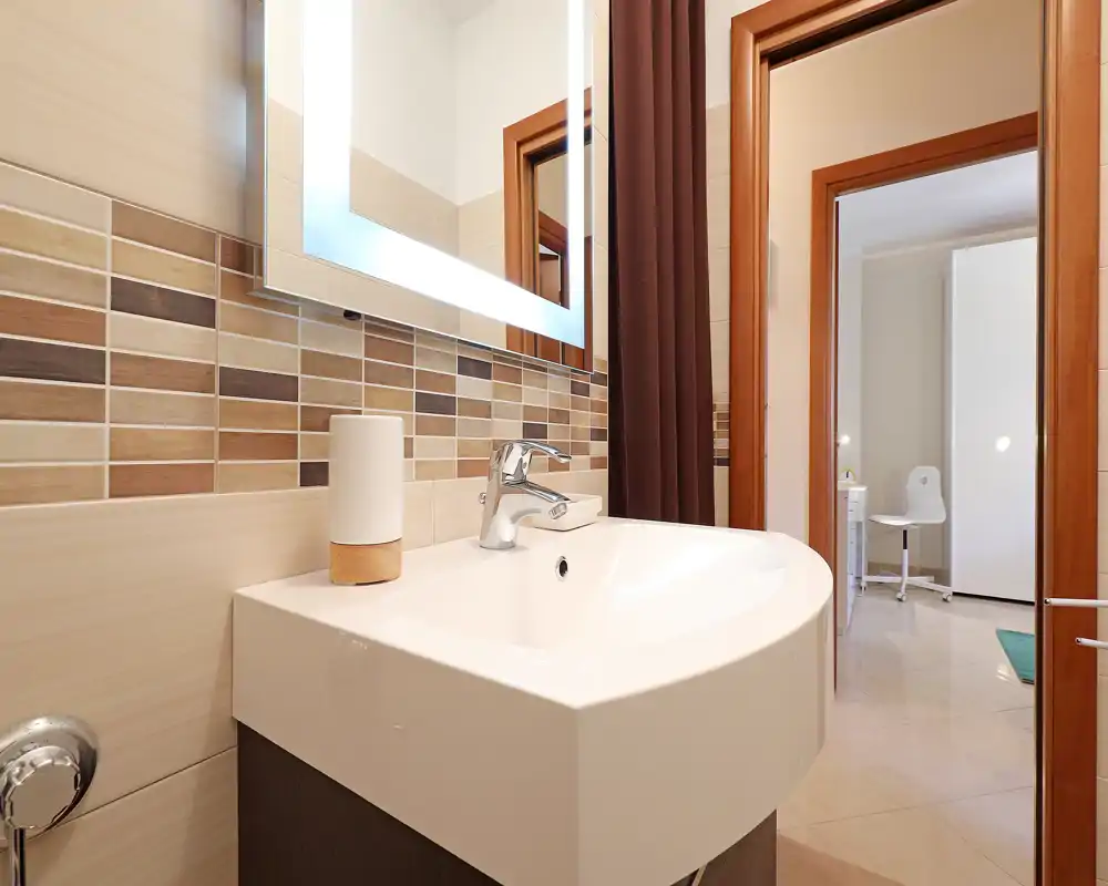 Baño moderno con lavabo blanco, grifo cromado y espejo iluminado; azulejos mosaico beige y marrones y puerta que comunica con una habitación contigua.