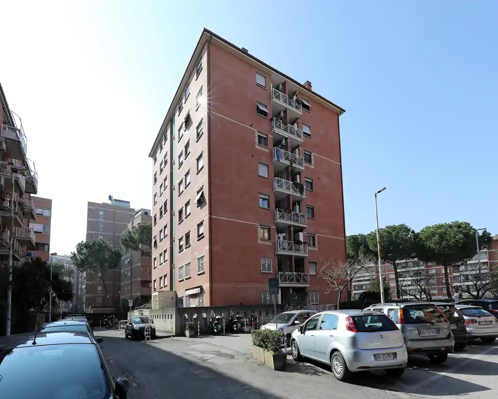 Vista exterior de un bloque residencial con coches aparcados y balcónes bajo un cielo despejado; adecuada para mostrar la fachada del edificio y la zona de aparcamiento.