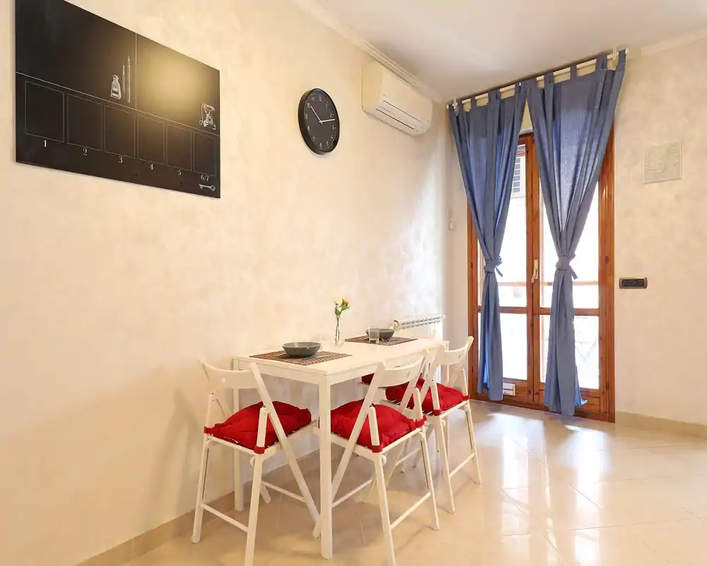 Zona de comedor luminosa y compacta con mesa blanca y cuatro sillas con cojines rojos, vajilla cuidada y un jarrón, junto a una puerta acristalada con cortinas azules que dejan pasar la luz natural.
