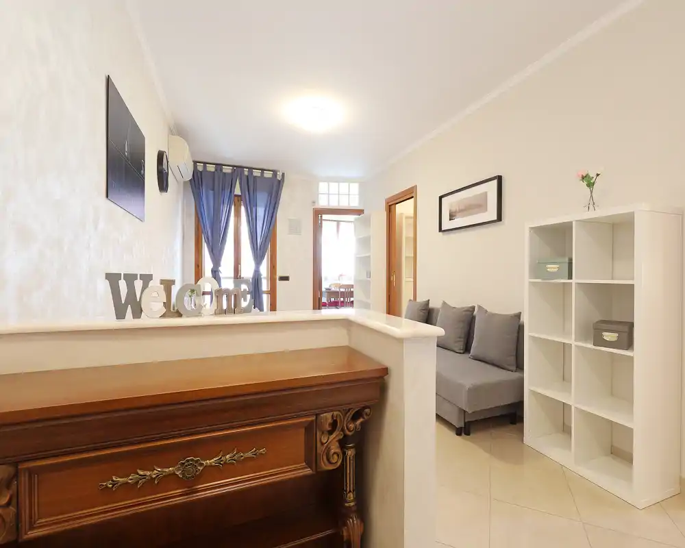 Sala de estar luminosa y ordenada con un sofá gris compacto, estantería decorativa y detalles acogedores; la luz natural a través de las puertas con cortinas aporta calidez.