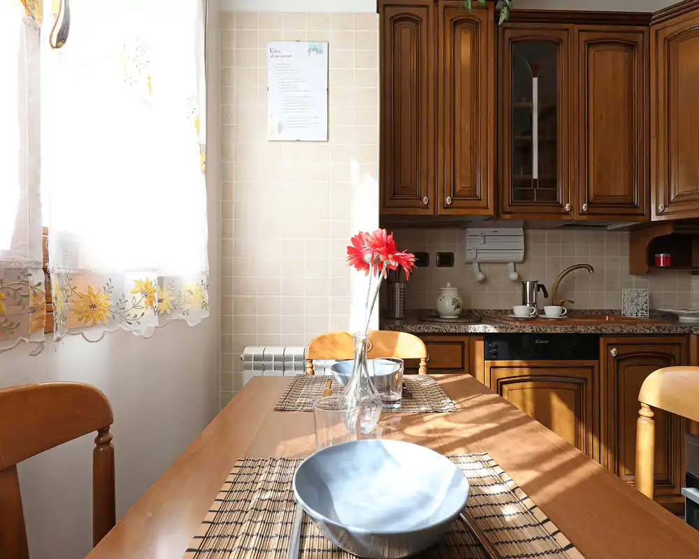 Cocina iluminada por el sol con cálidos muebles de madera y una pequeña mesa con individuales y un jarrón de flores rojas; espacio acogedor y luminoso para comidas informales.