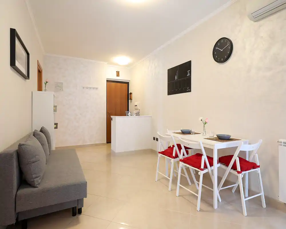 Sala/comedor moderno y luminoso con sofá compacto y mesa de comedor con sillas con cojines rojos. Tonalidades neutras y puesta en escena cuidada crean un espacio acogedor, ideal para alquileres de corta estancia.