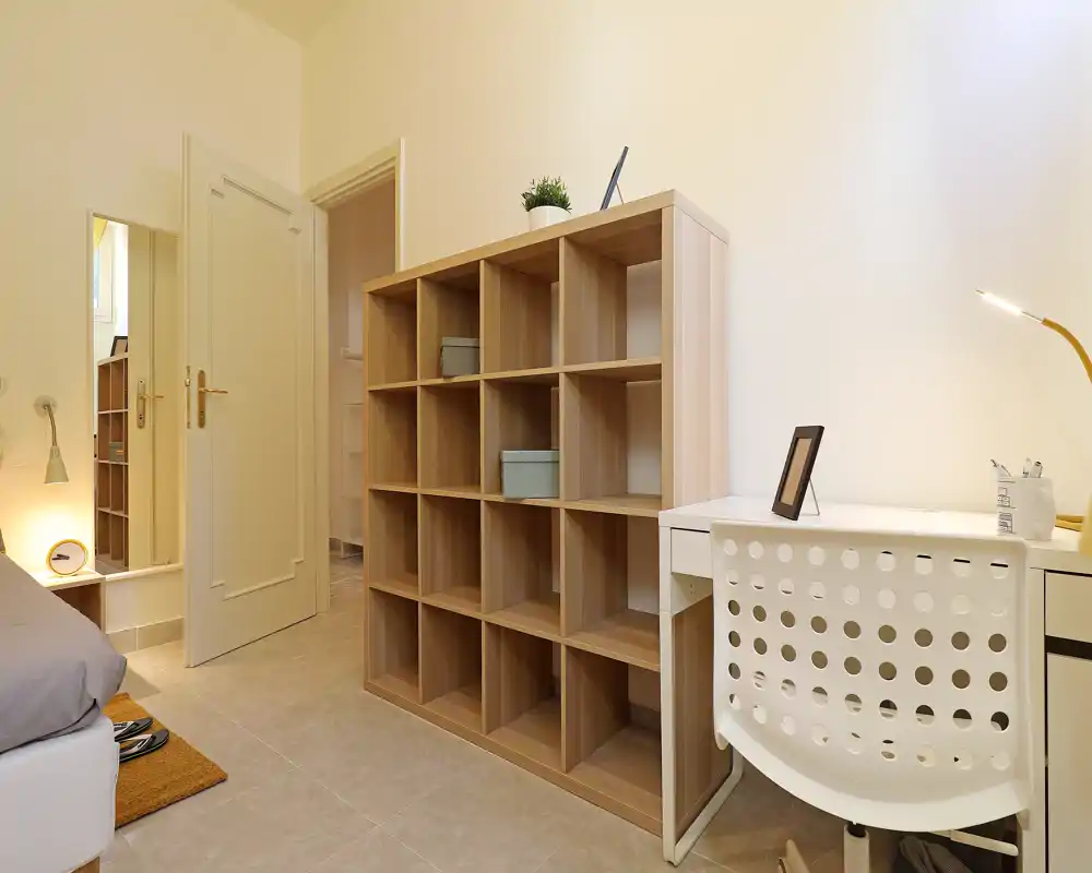 Bright, simply styled bedroom corner with a visible bed edge, a wooden cube shelving unit and a small desk — ideal for a single occupant seeking a tidy, functional room.