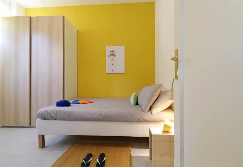 Bright, modern single bedroom with a comfortable bed against a warm yellow accent wall, bedside table with lamp, wardrobe and a small rug — ideal for a short-stay rental.