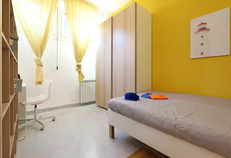 Sunny, compact bedroom with a single bed, wardrobe and a small desk by the window. Bright yellow accent wall and soft natural light create a cheerful, welcoming atmosphere ideal for a short-stay rental.
