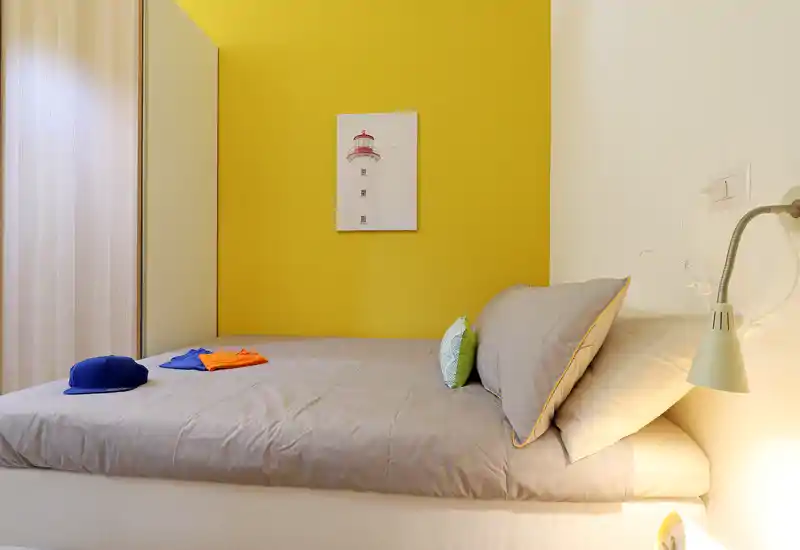 Bright, neatly staged bedroom with a grey bed against a cheerful yellow accent wall, bedside reading lamp and simple wall art.