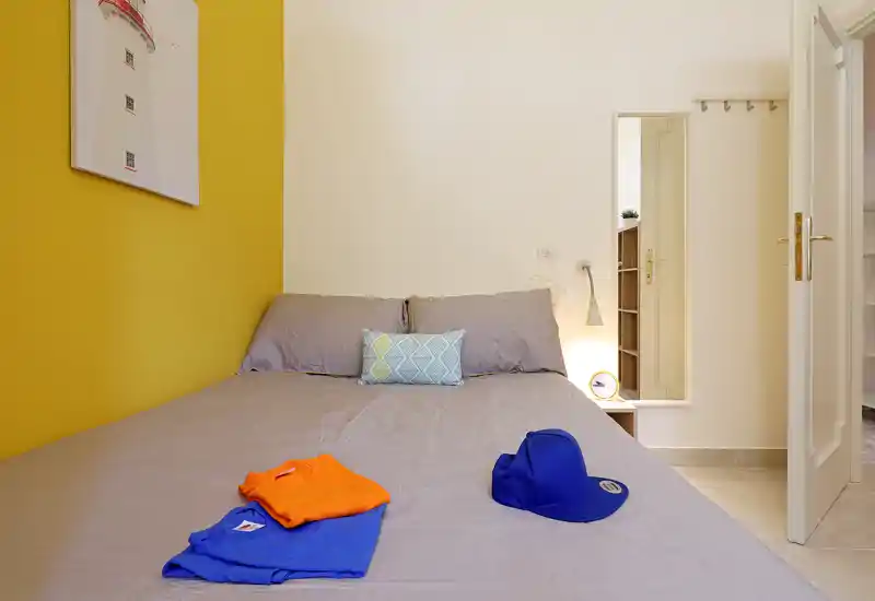 Bright, modern bedroom with a full bed against a warm yellow accent wall, decorative cushion and bedside lamp; neatly styled and well lit for listing photos.