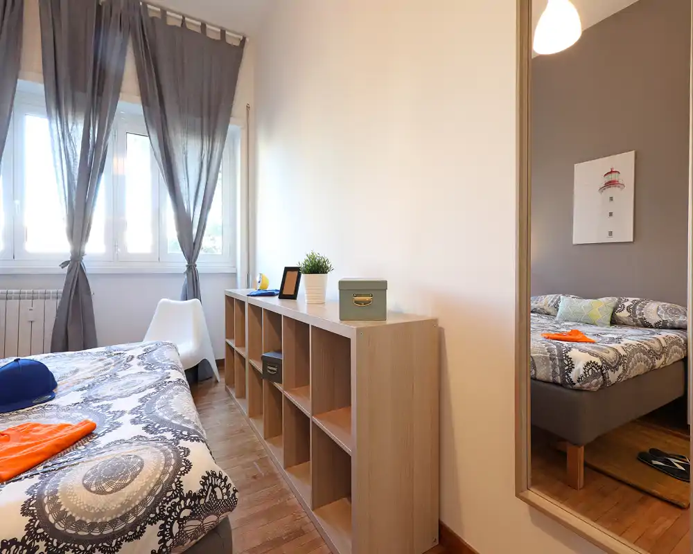 Dormitorio luminoso y ordenado con ropa de cama estampada, suelo de madera y una gran ventana que aporta luz natural. Un espejo de cuerpo entero refleja la cama y amplía la sensación de espacio.