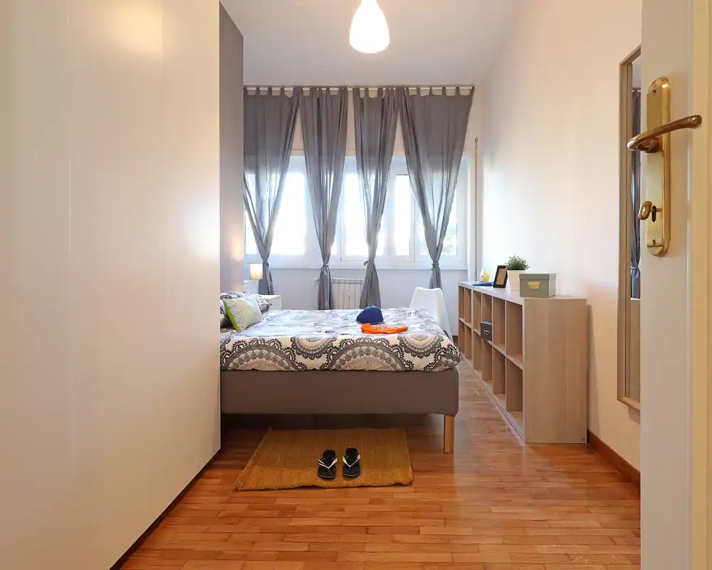 Dormitorio luminoso y ordenado con cama doble frente a grandes ventanas y cortinas grises; suelos de madera y estanterías discretas crean un ambiente moderno y relajado.