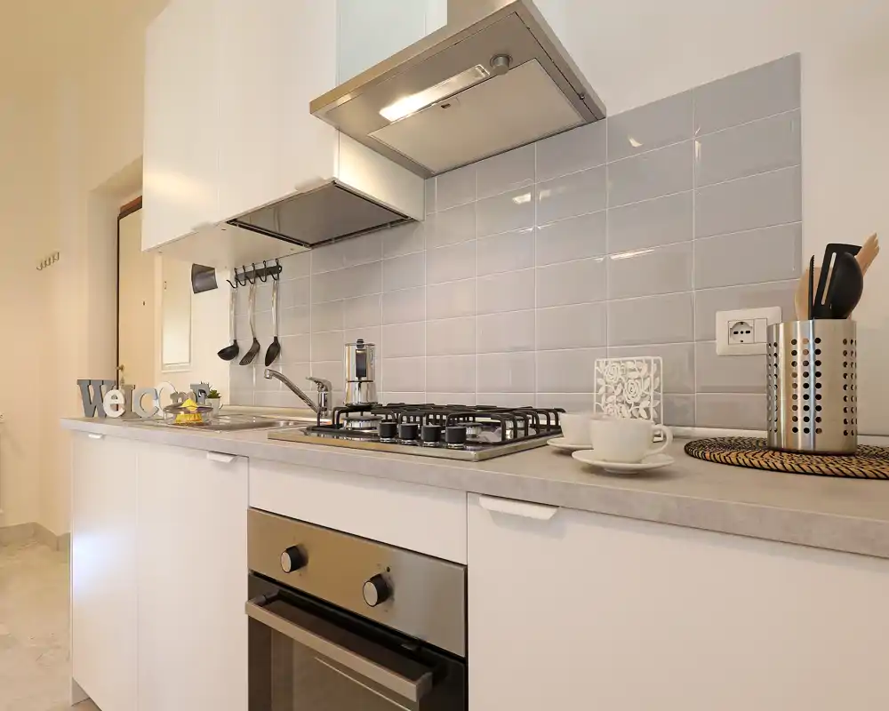 Modern compact kitchen with a gas hob, built-in oven and tiled backsplash. Styled with cups, utensils and a small welcome sign for a cozy, inviting look.