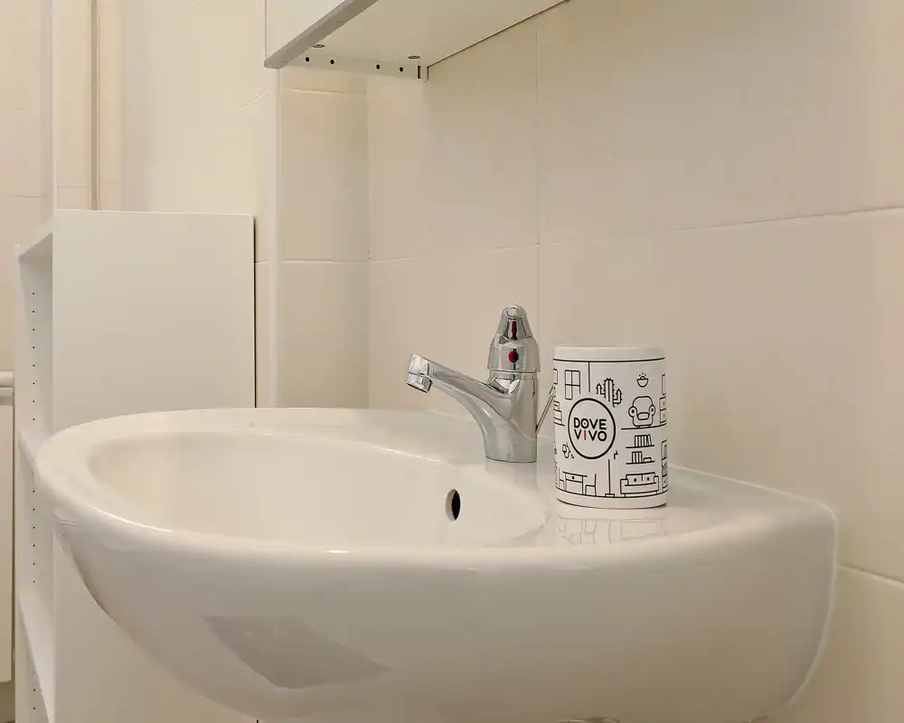 Clean bathroom sink with a modern single-handle faucet and a small accessory canister placed on the basin edge. Neutral tiled walls and a minimalist shelf are visible in the background.
