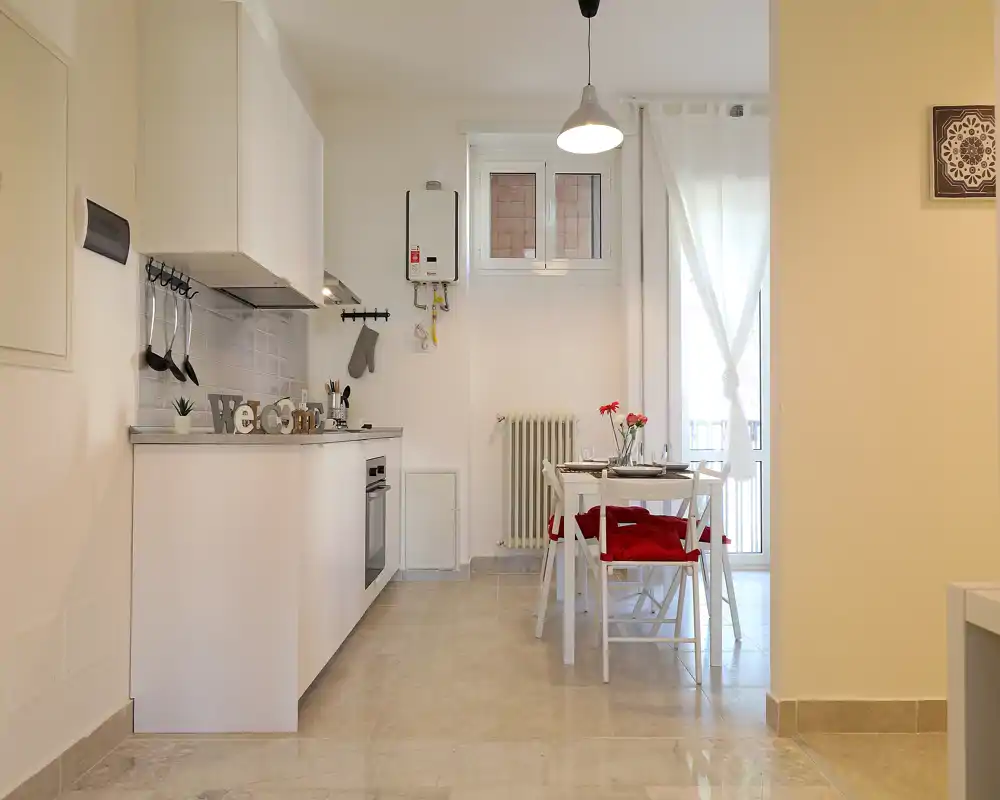Modern open kitchen with white cabinetry, built-in oven and a small dining table set by a bright window — clean, minimal and welcoming.