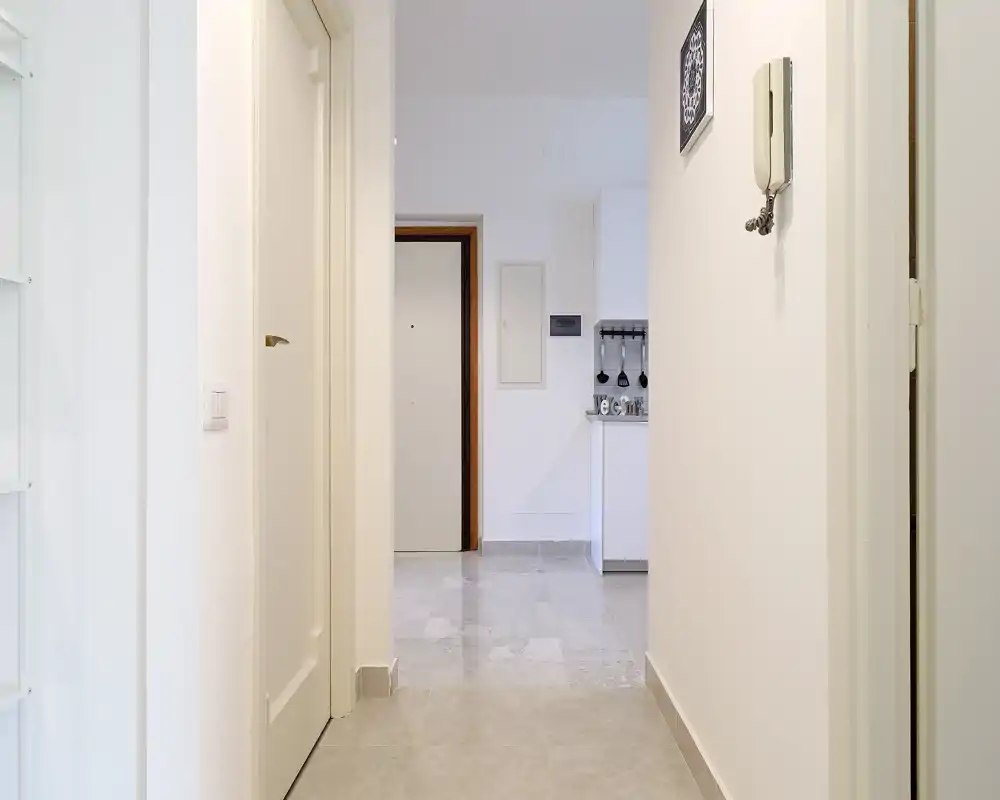 Bright, minimalist entrance corridor with neutral walls, tiled floor and a visible front door at the end of the hall.