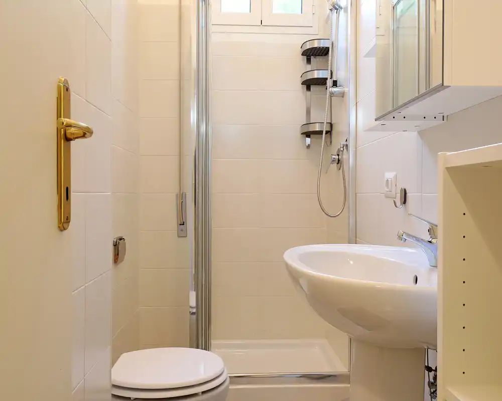 Compact, well-lit bathroom with a corner shower, pedestal sink and toilet; neutral tiles and chrome fixtures create a clean, modern look.