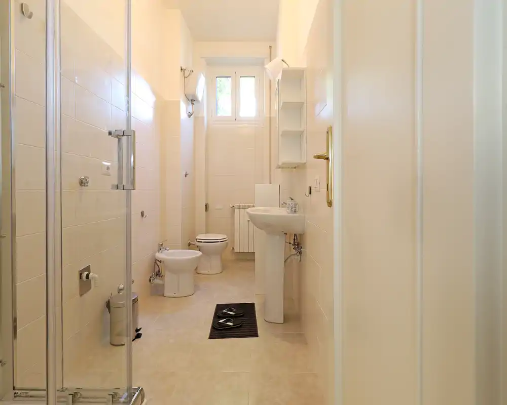 Bright, well-kept bathroom with a shower enclosure, bidet, toilet and pedestal sink. Neutral tiles and a high window provide natural light for a fresh, airy feel.