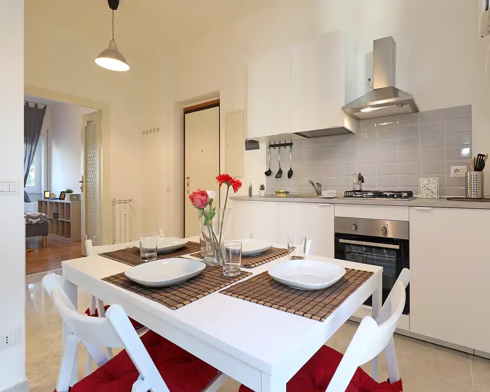 Bright, modern open kitchen with a set dining table for four. White cabinetry, stainless-steel oven and hood, and decorative placemats with fresh flowers create a welcoming, well-lit cooking and dining space.