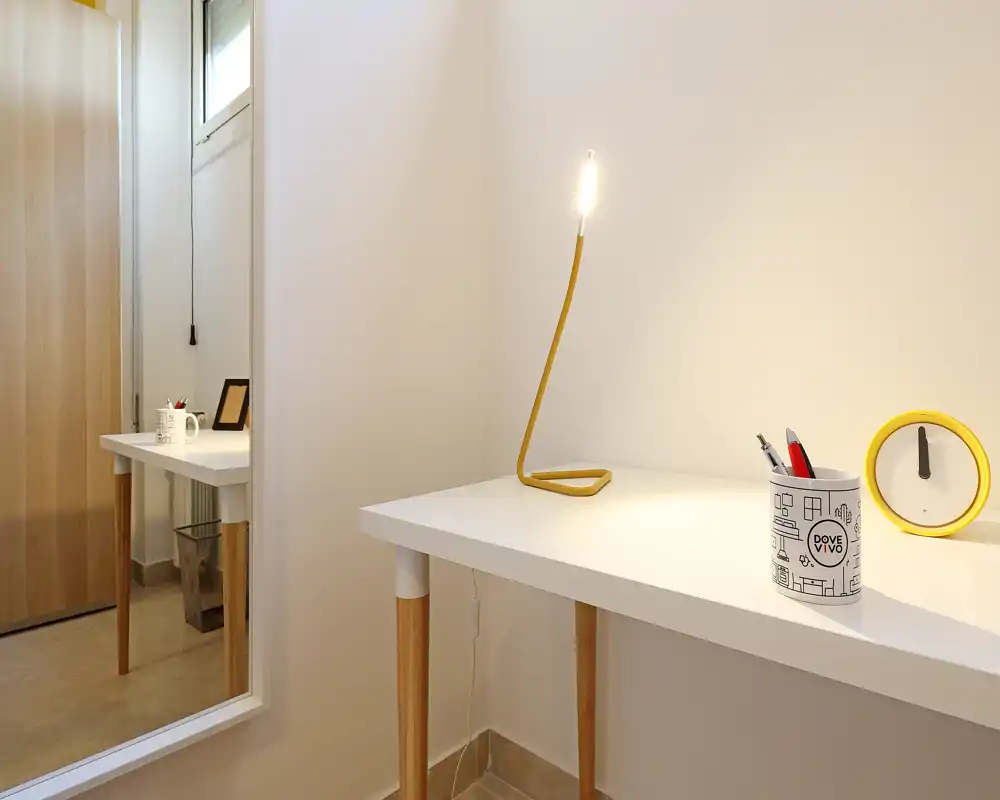 Angolo scrivania piccolo e ordinato con lampada da lettura gialla moderna, porta penne e una sveglia rotonda — ambiente luminoso e minimale contro una parete chiara.