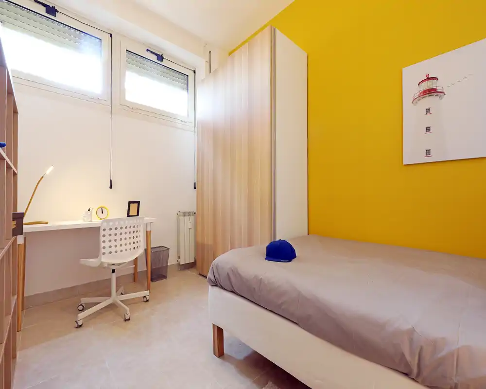 Camera singola luminosa e moderna con letto ordinato, scrivania sotto finestre alte e grande armadio contro una parete gialla vivace.