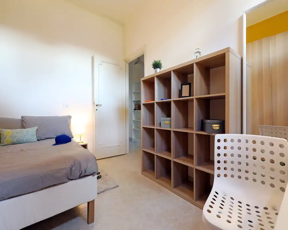 Camera da letto luminosa e semplice, con letto rifatto, lampada da comodino e scaffalatura aperta che offre spazio e arredo sobrio.