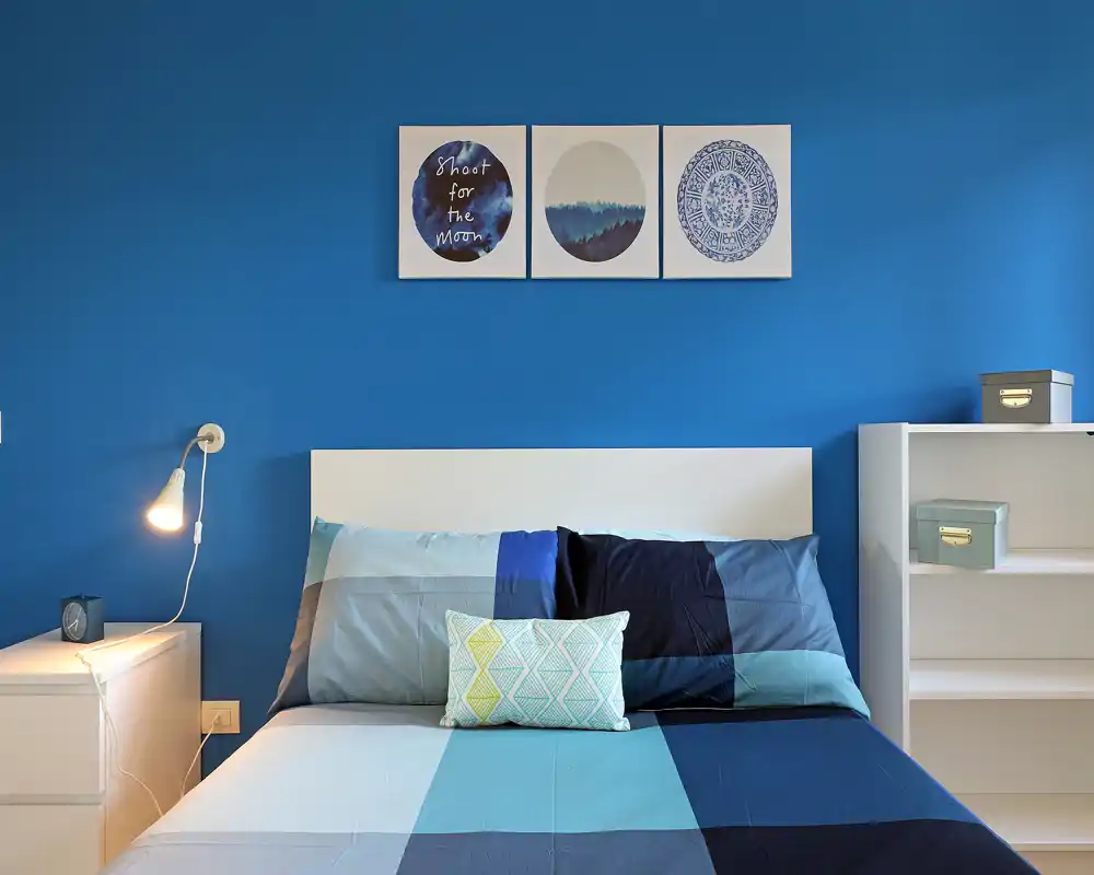 Dormitorio moderno y luminoso con cama doble bien hecha en tonos azules, cojines decorativos y cuadros coordinados; líneas limpias y una lámpara de noche que aportan una atmósfera serena y con estilo.