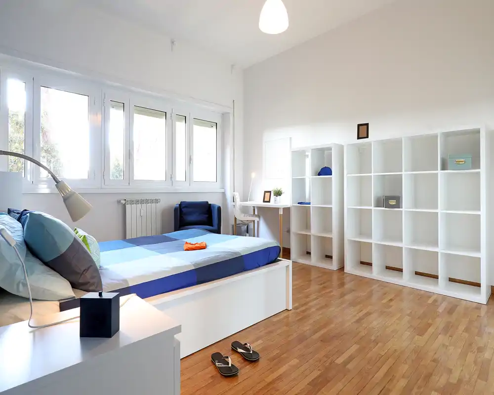 Dormitorio moderno y luminoso con una cama grande hecha, ventanas amplias y estanterías minimalistas: ideal para anuncios que destaquen el espacio y la luz natural.