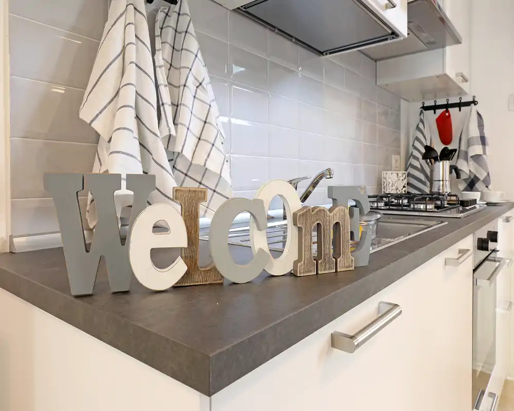 Encimera de cocina moderna y luminosa con un adorno "welcome", salpicadero de azulejos, fregadero y placa de gas — una cocina cuidada y acogedora ideal para alquileres vacacionales.