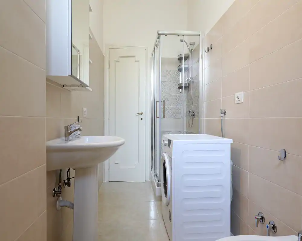 Baño moderno y luminoso con cabina de ducha de cristal, lavabo de pedestal y una lavadora situada junto a la ducha.