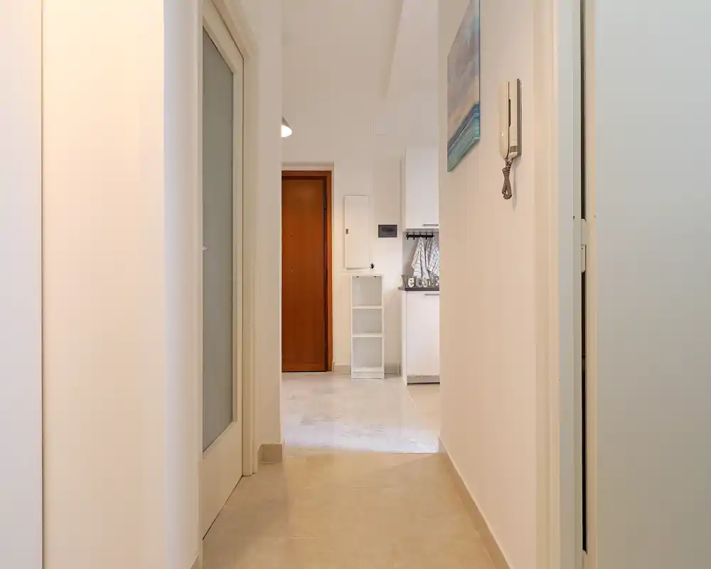 Entrada bien iluminada con suelo de baldosas, puerta principal e interfono visibles: un área de acceso limpia y neutra que conduce al apartamento.