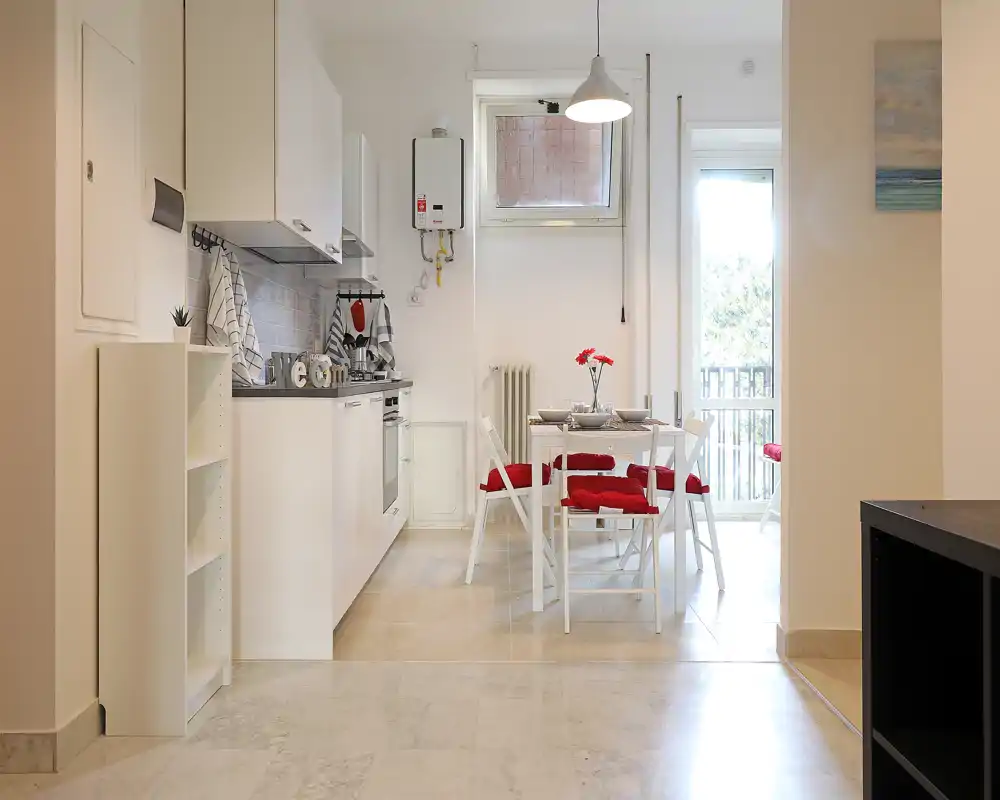 Cocina moderna y luminosa en espacio abierto con una pequeña mesa de comedor con cojines rojos y puerta al balcón, ideal para apartamentos urbanos compactos.