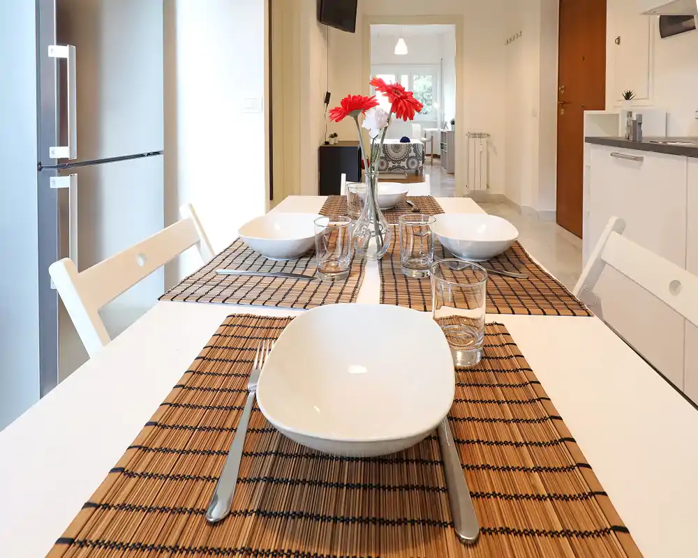 Área de comedor moderna y luminosa con mesa puesta para cuatro, individuales decorativos y flores; vista clara hacia la sala contigua.
