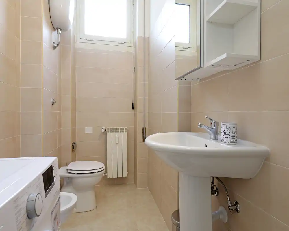 Baño compacto y moderno con lavabo de pedestal, inodoro, armario con espejo y lavadora. Azulejos beige neutro y luz natural brillante que crean un espacio limpio y funcional.