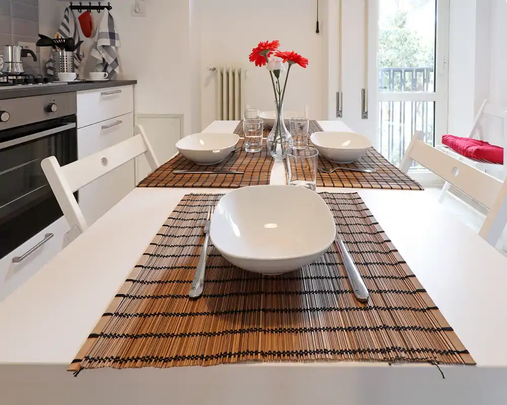 Zona de comedor moderna y luminosa con mesa blanca puesta, manteles individuales y un pequeño jarrón con flores rojas junto a la cocina y la puerta al balcón.