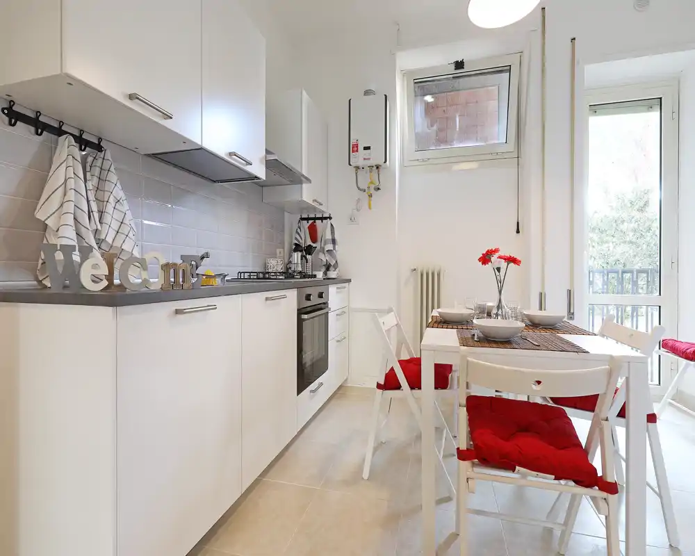 Cocina moderna y luminosa con muebles blancos, horno integrado y una mesa de comedor pequeña con cojines rojos y flores decorativas — ideal para anuncios de apartamentos compactos.
