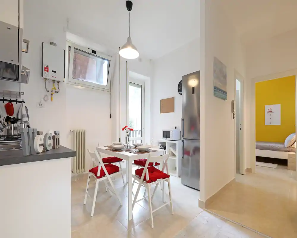 Zona de comedor luminosa y moderna en cocina abierta con mesa cuadrada para cuatro y cojines rojos llamativos, perfecta para comidas informales.
