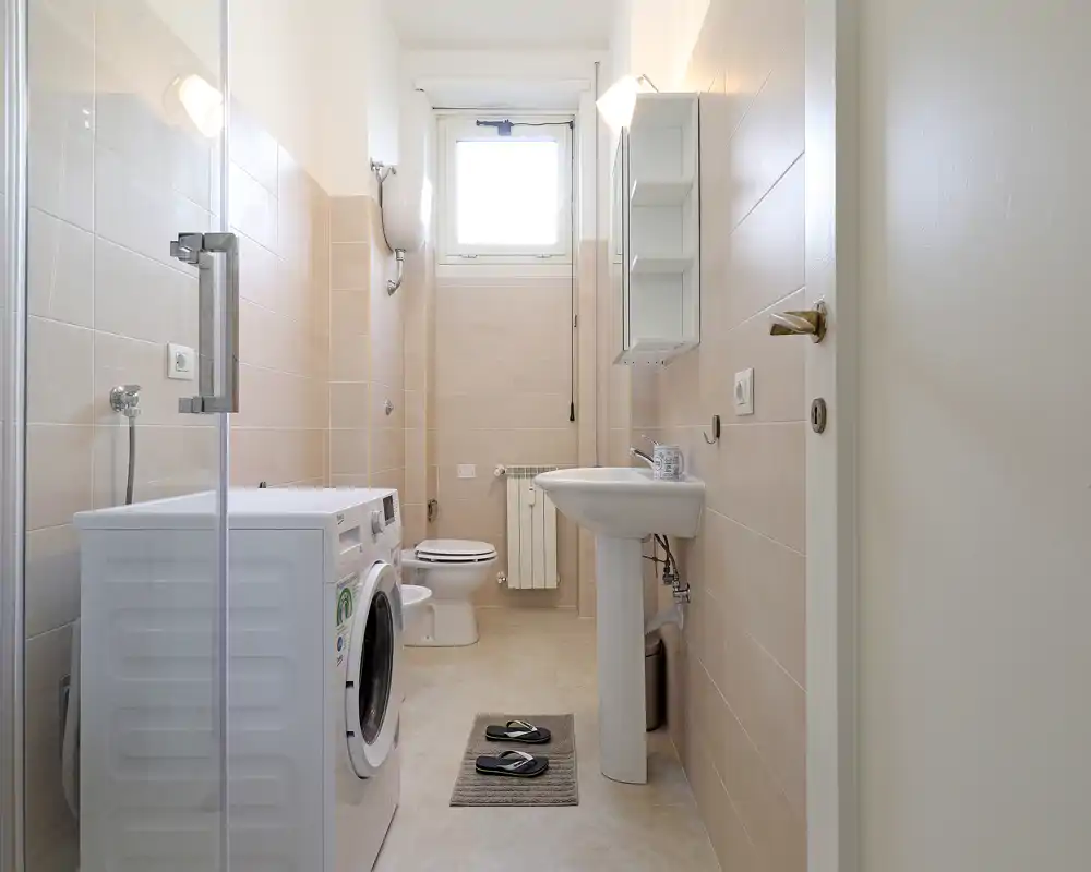 Baño luminoso y bien cuidado con lavabo de pedestal, inodoro, cabina de ducha y lavadora — azulejos neutros y una ventana alta que proporciona buena luz natural.