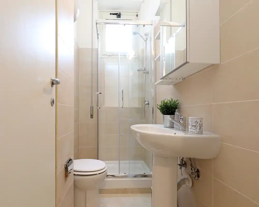 Baño compacto y bien iluminado con ducha acristalada, lavabo de pedestal y inodoro. Azulejos de tono neutro y mueble con espejo para una sensación limpia y moderna.