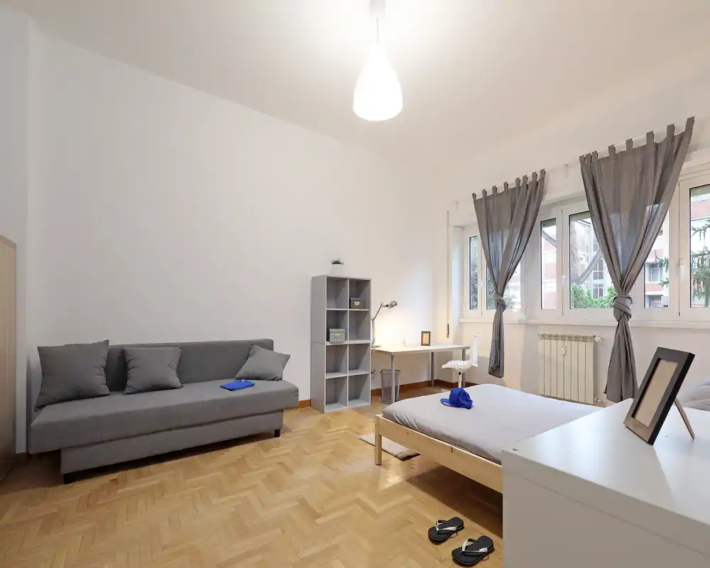 Bright, minimalist bedroom with a single bed, desk by the window and a grey sofa. Natural light, neutral tones and parquet flooring create a calm, modern atmosphere ideal for renters.