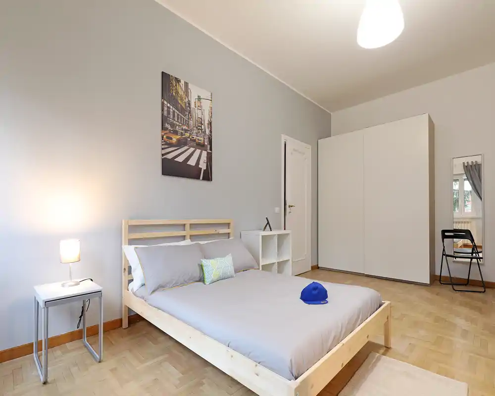 Bright, modern bedroom with a minimalist wooden double bed, soft grey bedding, a bedside table with lamp and a large white wardrobe — calm neutral tones and clean lines.