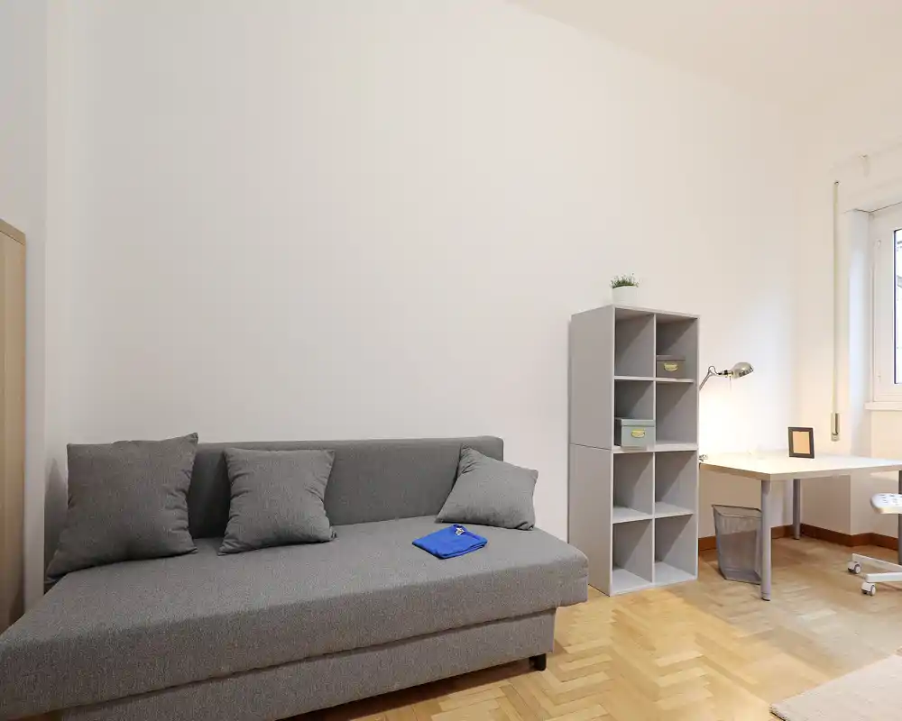 Minimal, well-lit living area with a grey sofa, cushions, shelving unit and a small desk by the window — bright and uncluttered space ideal for relaxing or working.