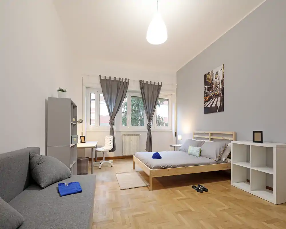 Bright, modern bedroom with a wooden bed frame, neutral linens and a cozy seating area by the window. Large windows with sheer curtains, a small desk and minimalist shelving make the space functional and airy.