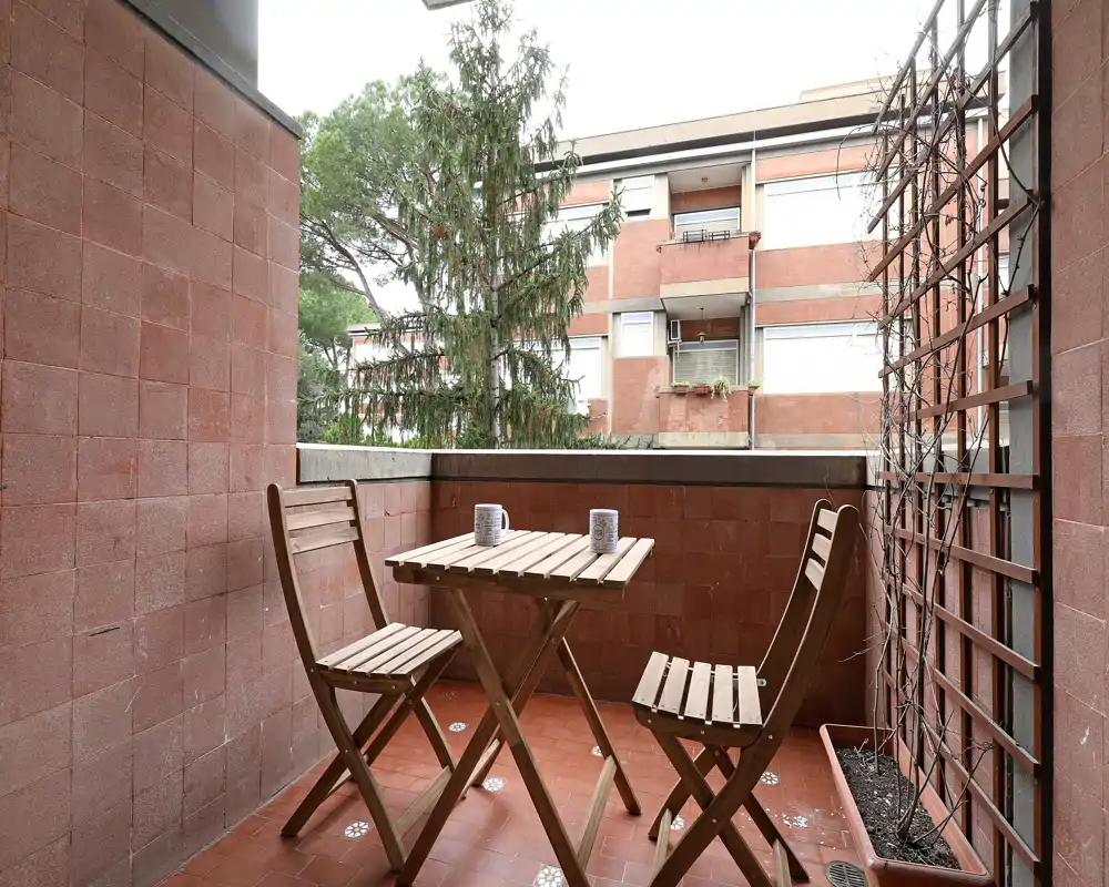 Sunny small balcony with a wooden bistro table and two folding chairs, ideal for morning coffee. The space is neatly staged with two mugs on the table and a trellis planter at the side.