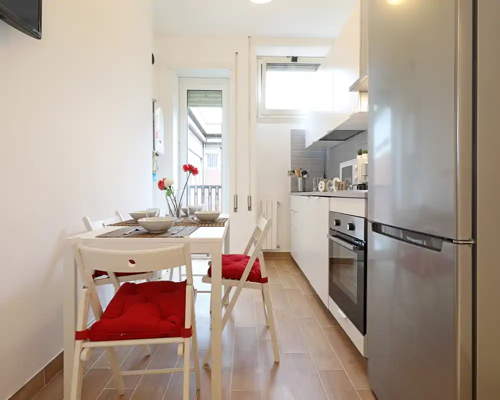 Compact modern kitchen with white cabinets, stainless fridge and built-in oven. A small dining table with red-cushioned chairs and a vase of flowers gives a cozy, well-lit feel.