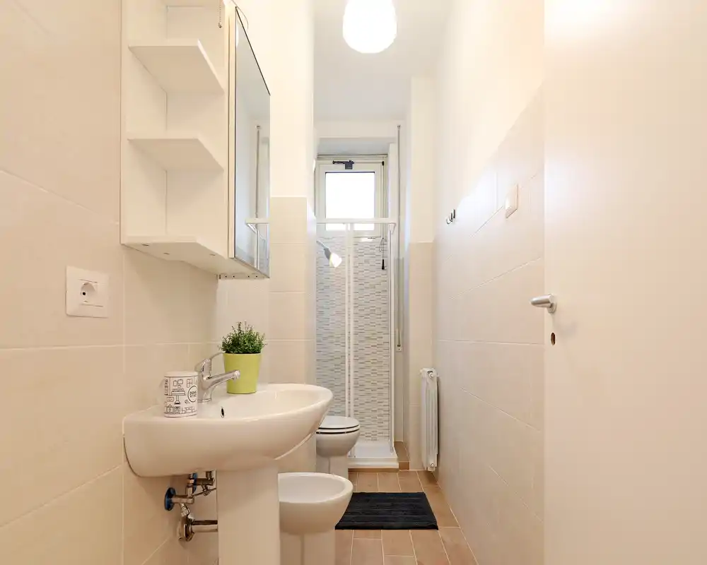 Bright, modern bathroom with a pedestal sink, bidet, toilet and a compact shower cubicle at the far end. Neutral tiles and simple white fixtures create a clean, minimalist look.