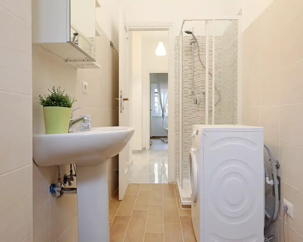 Bright, modern bathroom with a pedestal sink, shower enclosure and a washing machine; neutral tiles and wood-effect floor create a clean, functional space.